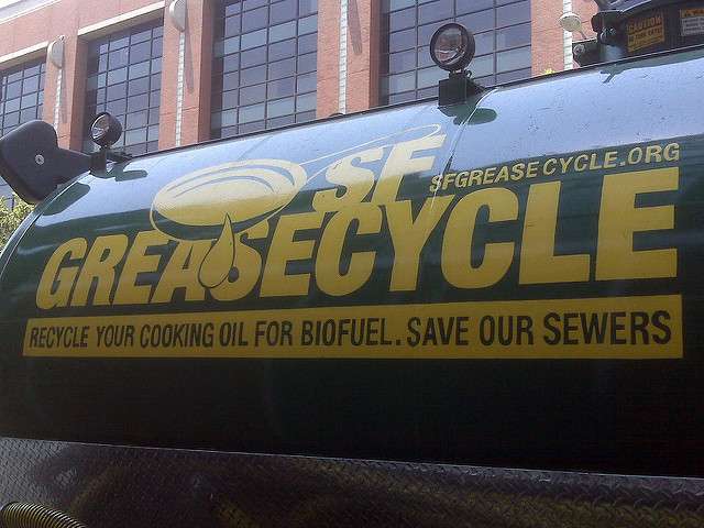 A green truck with “SF Greasecycle” and “Recycle your cooking oil for biofuel. Save our sewers” printed on the side, highlighting grease trap recycling, is parked in front of a brick building.