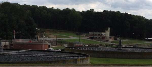 A wastewater treatment facility with several circular tanks and grease trap systems in the foreground, grassy areas, trees, and buildings in the background under a cloudy sky.