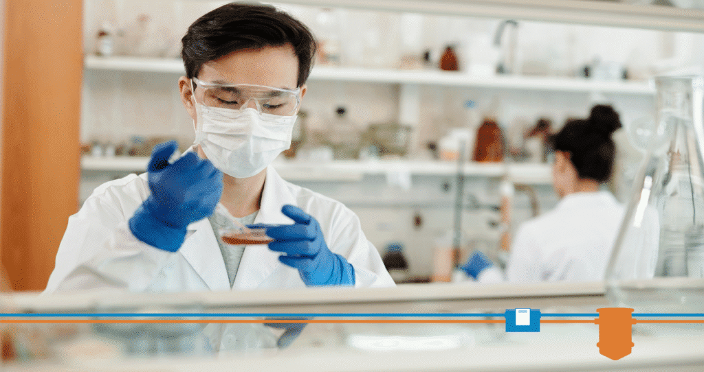 A person wearing safety goggles, a mask, and blue gloves holds a Petri dish and uses a pipette in a laboratory. Shelves with lab equipment, including grease trap testing supplies, and another person working are visible in the background.