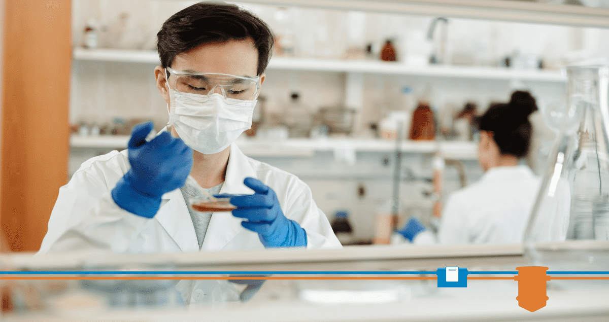 A person wearing safety goggles, a mask, and blue gloves holds a Petri dish and uses a pipette in a laboratory. Shelves with lab equipment, including grease trap testing supplies, and another person working are visible in the background.