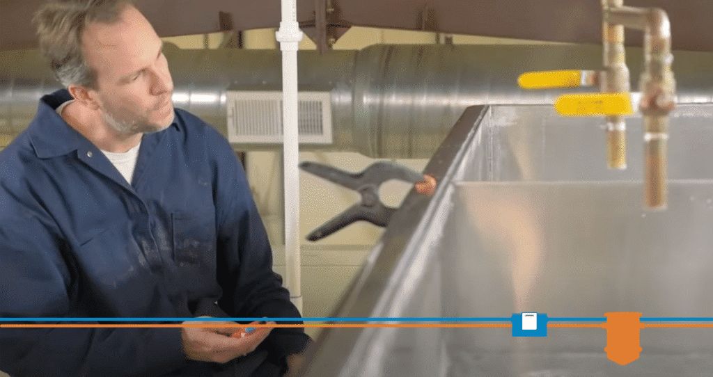 A man in a blue work uniform inspects a metal tank with attached water pipes, a clamp, and a grease trap. Industrial pipes and ventilation ductwork are visible in the background.