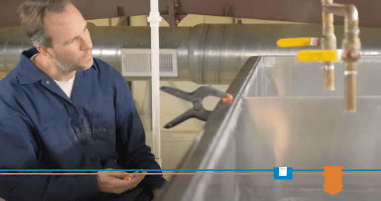 A man in a blue work uniform inspects a metal tank with attached water pipes, a clamp, and a grease trap. Industrial pipes and ventilation ductwork are visible in the background.