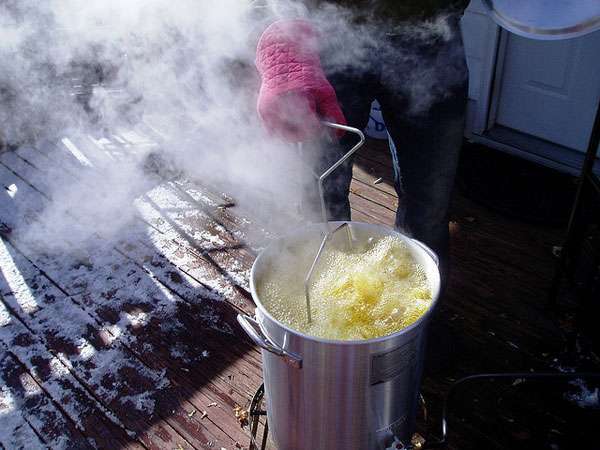 A person wearing a pink oven mitt lowers an item into a large metal pot of hot oil, which bubbles and steams beside a grease trap on a wooden outdoor deck with patches of snow visible.