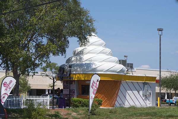 A small ice cream shop shaped like a giant soft-serve cone with a white swirled top and orange base, featuring a modern grease trap, sits near trees and a parking lot on a sunny day.