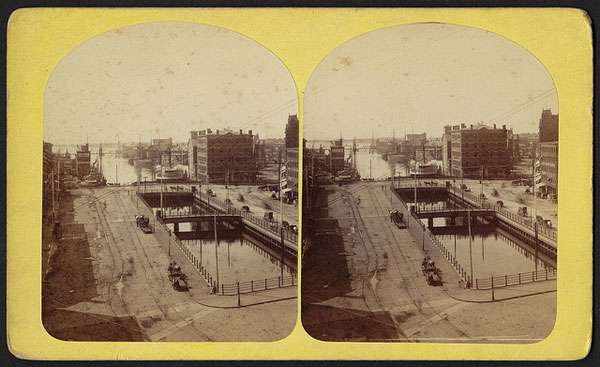 A stereoscopic image shows a street with horse-drawn carriages and a canal—possibly channeling runoff to a grease trap—bordered by multi-story brick buildings and industrial structures near a waterfront.