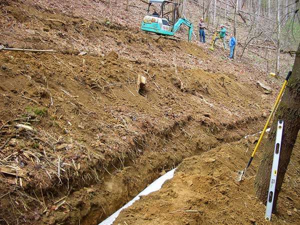 A long, narrow trench is dug along a hillside in a wooded area, likely for a grease trap installation. An excavator is parked nearby while three people above use surveying tools. A measuring rod and level are positioned by the trench.