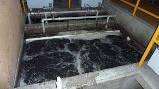 A rectangular concrete tank, likely a grease trap, filled with dark, bubbling water and surrounded by pipes, railings, and equipment—typical of an industrial or wastewater treatment facility.