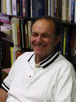 A man wearing a white polo shirt with black stripes on the collar is smiling while seated in front of a bookshelf filled with books and binders, possibly discussing topics like maintenance or grease trap solutions.