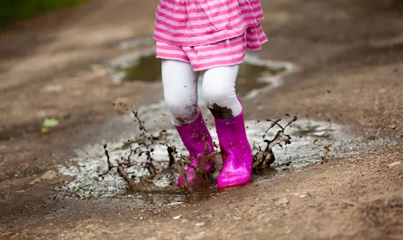 A child wearing a pink and white striped dress, white tights, and bright pink rain boots jumps in a muddy puddle near a grease trap, causing mud and water to splash up.