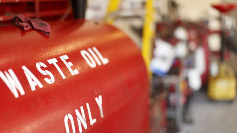 A red tank labeled "WASTE OIL ONLY" stands out in white letters. The blurred background reveals indistinct objects and equipment, hinting at an industrial setting with potential grease trap maintenance nearby.